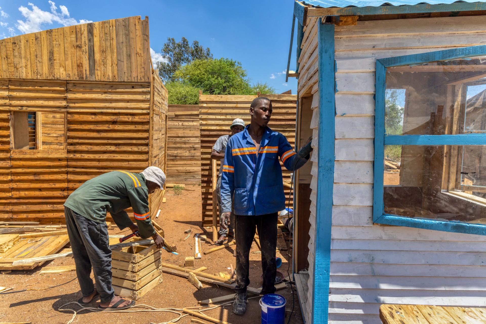 three african american workers making a wendy house, wooden shed outdoors, they hammering pallets to make planks and assemble them in panels